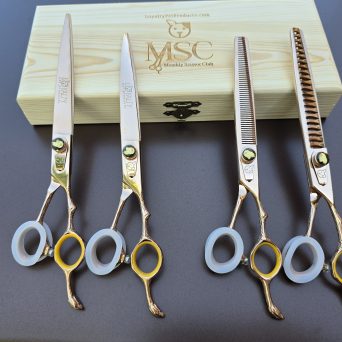 A photo of a 4 pc golden set of grooming shears laid on a wooden box with a black background